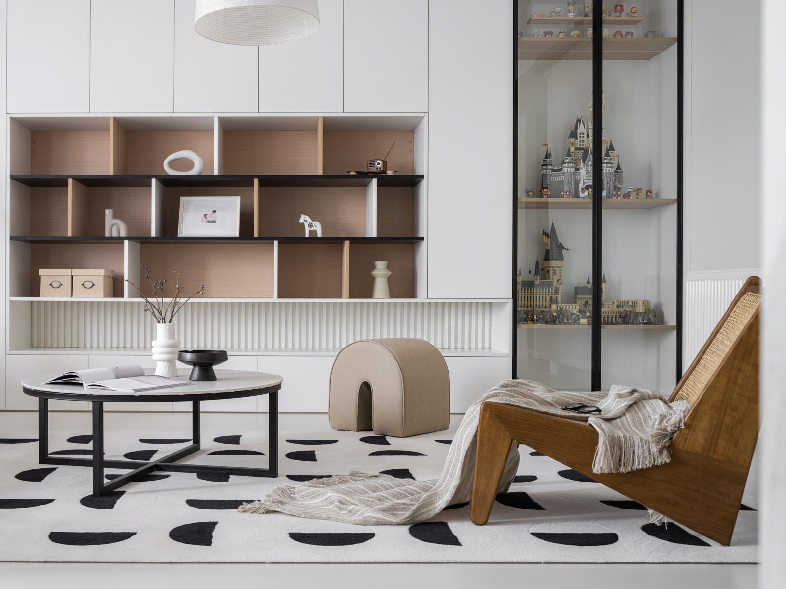Living room with white built-in cabinetry, oak-lined shelving, steel-framed glass display cabinet, spotted rug and cane lounge chair